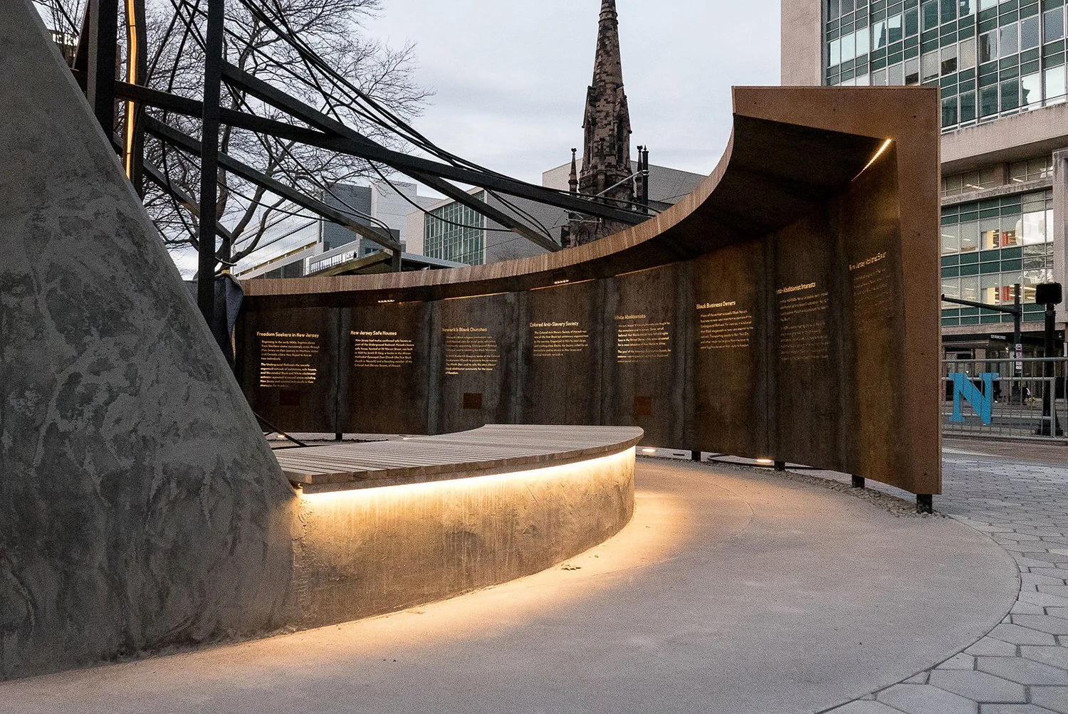 Inside the completed monument: cut text in a curved steel wall, black locust seating and warm light