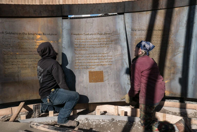 Two people fitting the Freedom Seekers in New Jersey steel panel to the learning wall
