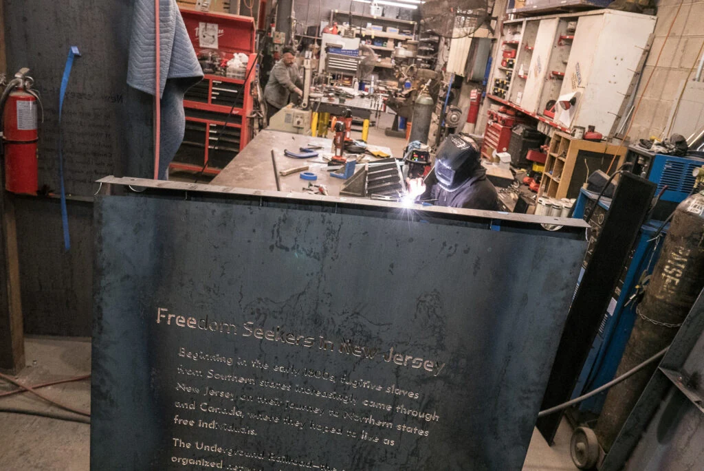 A welder working behind a steel panel headed Freedom Seekers in New Jersey